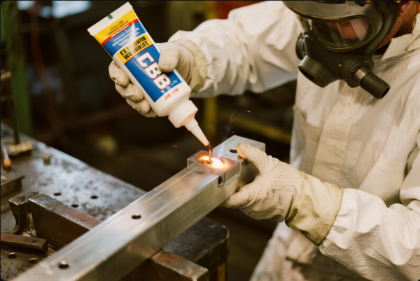 A technician in full protective clothing and a respirator mask applies a specialized welding compound labeled "CBB" to a metal workpiece that is being heated, as sparks indicate active bonding. The process takes place in a workshop setting with a metal table and tools visible in the background. This scene showcases advanced manual welding or brazing techniques for precision metal joining in an industrial environment.