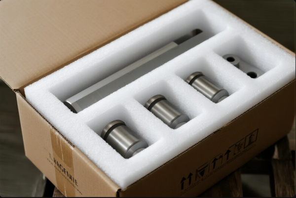 The image displays a set of cylindrical metal parts neatly arranged in a cardboard shipping box. The components are individually separated and cushioned by custom-cut white foam inserts, ensuring each part is protected from damage during transportation. The packaging highlights careful handling and a focus on quality assurance in the shipment of industrial components.