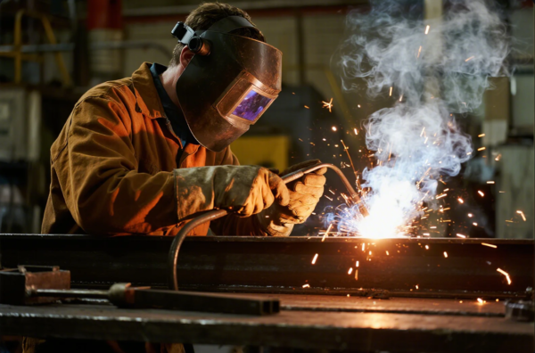This image shows a welder in a workshop, equipped with a dark welding helmet and heavy-duty gloves, using an arc welding torch to join metal pieces. Bright sparks and a large plume of smoke are visible at the welding point, highlighting the intensity and high temperature of the process. The welder's posture and focus emphasize precision and safety, and the industrial background suggests a professional metal fabrication environment.