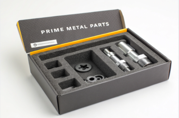 This image shows a high-quality presentation box with the label "PRIME METAL PARTS" on the inside of the lid. Inside, several precision-machined metal parts—including threaded shafts, a die, and small round components—are organized in custom-cut foam for secure storage and display. The box and packaging highlight attention to detail, quality control, and professionalism in delivering industrial or mechanical components.