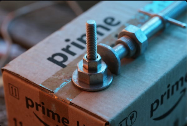 A macro shot showing a metallic bolt, nut, and washer assembly resting on a cardboard box labeled "Prime." The hardware is illuminated with a blend of blue and warm lighting, highlighting the precision threading and industrial texture. The background suggests a shipping or workshop environment, combining logistics branding with hardware manufacturing focus.