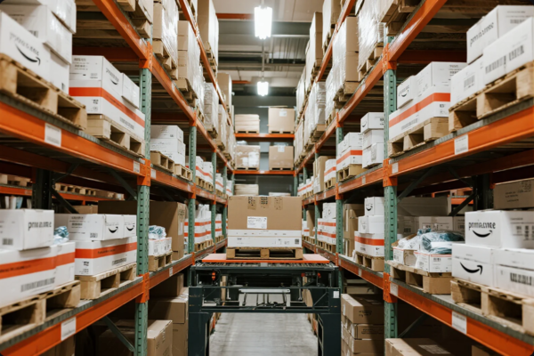 A clean and well-lit warehouse featuring multiple rows of tall industrial shelving loaded with neatly stacked Prime-branded cardboard boxes and pallets. A conveyor system is positioned in the center aisle, supporting the efficient movement and distribution of goods. The image showcases an organized inventory management system, ready for global order fulfillment.
