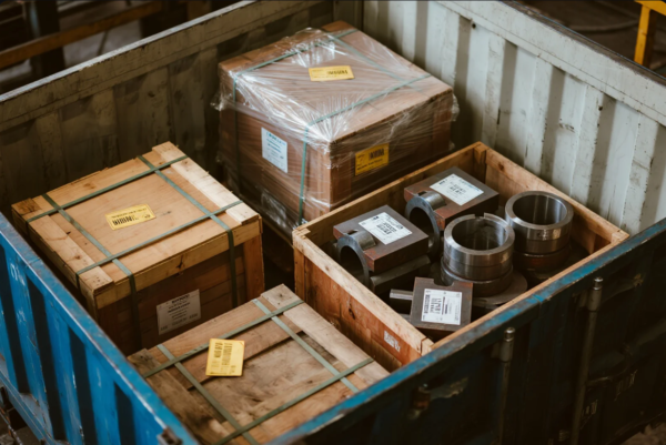 A steel shipping container holds several neatly arranged wooden crates, some sealed with plastic wrap and others open to reveal heavy metal components such as machined rings and square flanges. Each item is labeled with tracking and specification stickers, indicating careful organization and preparation for international shipment from a manufacturing facility.