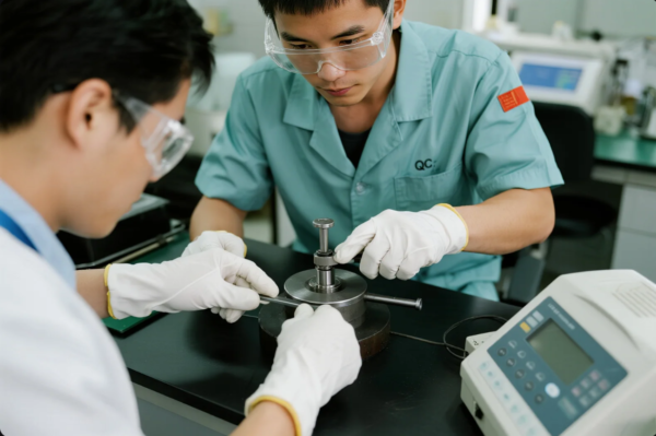 Two quality control engineers wearing safety goggles and gloves are conducting a precise measurement on a machined metal part using specialized equipment in a clean laboratory environment. One technician adjusts the alignment while the other records data, highlighting the factory’s commitment to strict quality assurance standards.