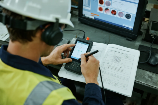 This image shows a factory quality control inspector in safety gear (white hard hat, yellow vest, and headphones) seated at a workstation. The inspector is operating a handheld electronic device with a stylus while reviewing data in a technical report or inspection record. A computer monitor nearby displays detailed inspection images and analysis results, indicating a thorough and modern approach to quality assurance and inspection in an industrial setting.