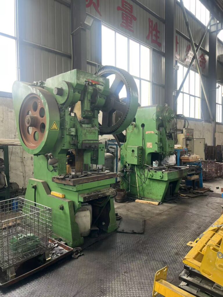 The image shows two large green mechanical stamping or punching machines positioned side by side inside a well-lit metalworking factory. The machines are equipped with large flywheels and worktables, and appear to be used for shaping or cutting metal parts. The factory environment features high ceilings, large windows, industrial flooring, and various tools and materials organized around the workspace. Chinese characters are visible on the upper wall, indicating the factory’s location or slogan. The atmosphere conveys a sense of heavy-duty manufacturing and precision engineering.