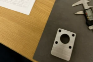 Machined metal bracket on inspection mat with digital caliper and QC sheet for dimensional verification.