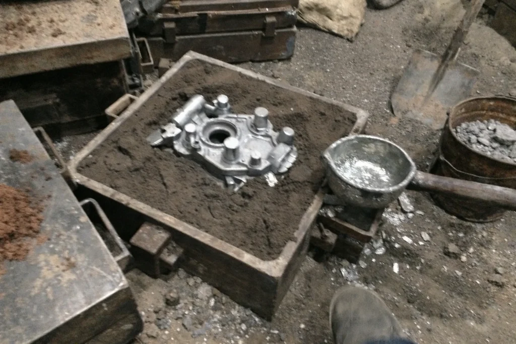 Inside the foundry workshop, custom metal sand casting parts, sand mold boxes, casting patterns, and scattered tools are visible.